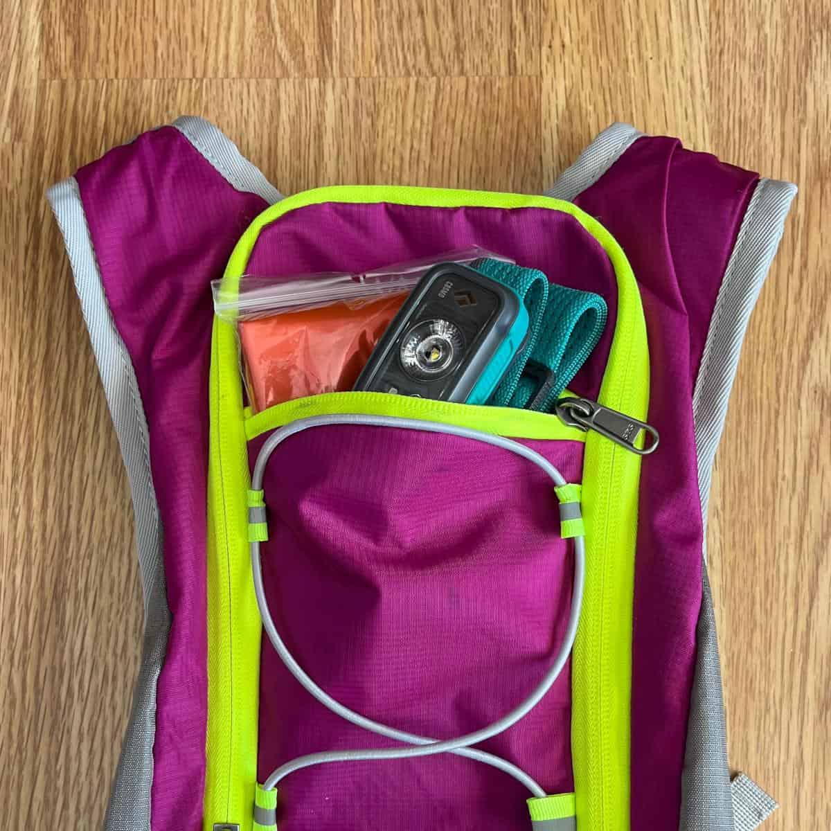 An orange emergency blanket and a teal headlamp poking out of the front pocket of a pink hiking backpack.