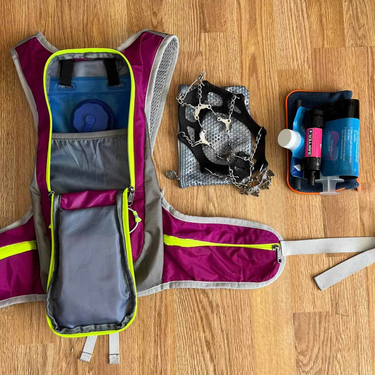 A pink hiking backpack with the main pocket opened, revealing a water bladder tucked inside. Next to the backpack is a pair of shoe spikes and a water filtration straw.