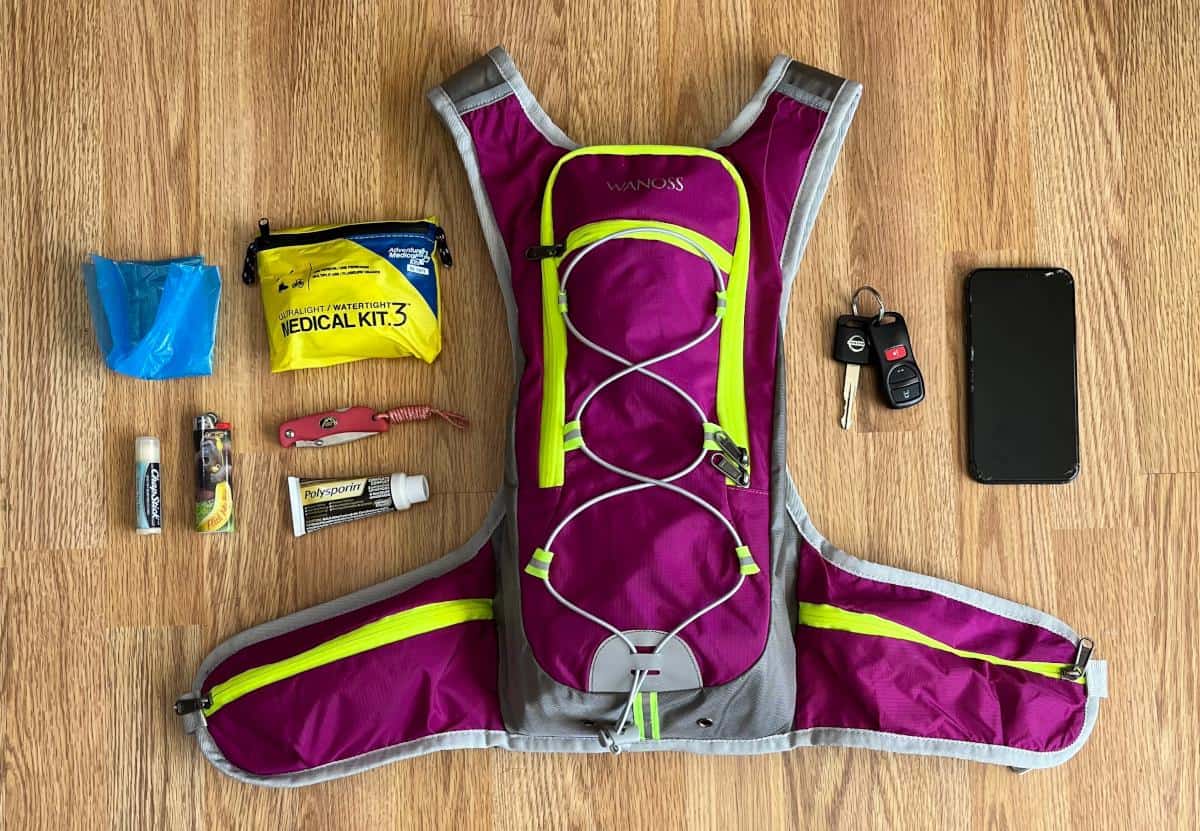 A pink hiking backpack laying on the floor. On one side of the backpack there is a Ziploc bag, a small first aid kit, a tube of Chapstick, a lighter, a pocket knife and a tube of Polysporin. On the other side of the backpack is a set of car keys and an iPhone.