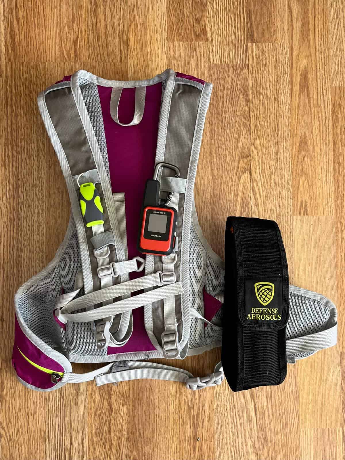 A whistle, a satellite communicator, and a canister of bear spray attached to the straps of a pink hiking daypack.