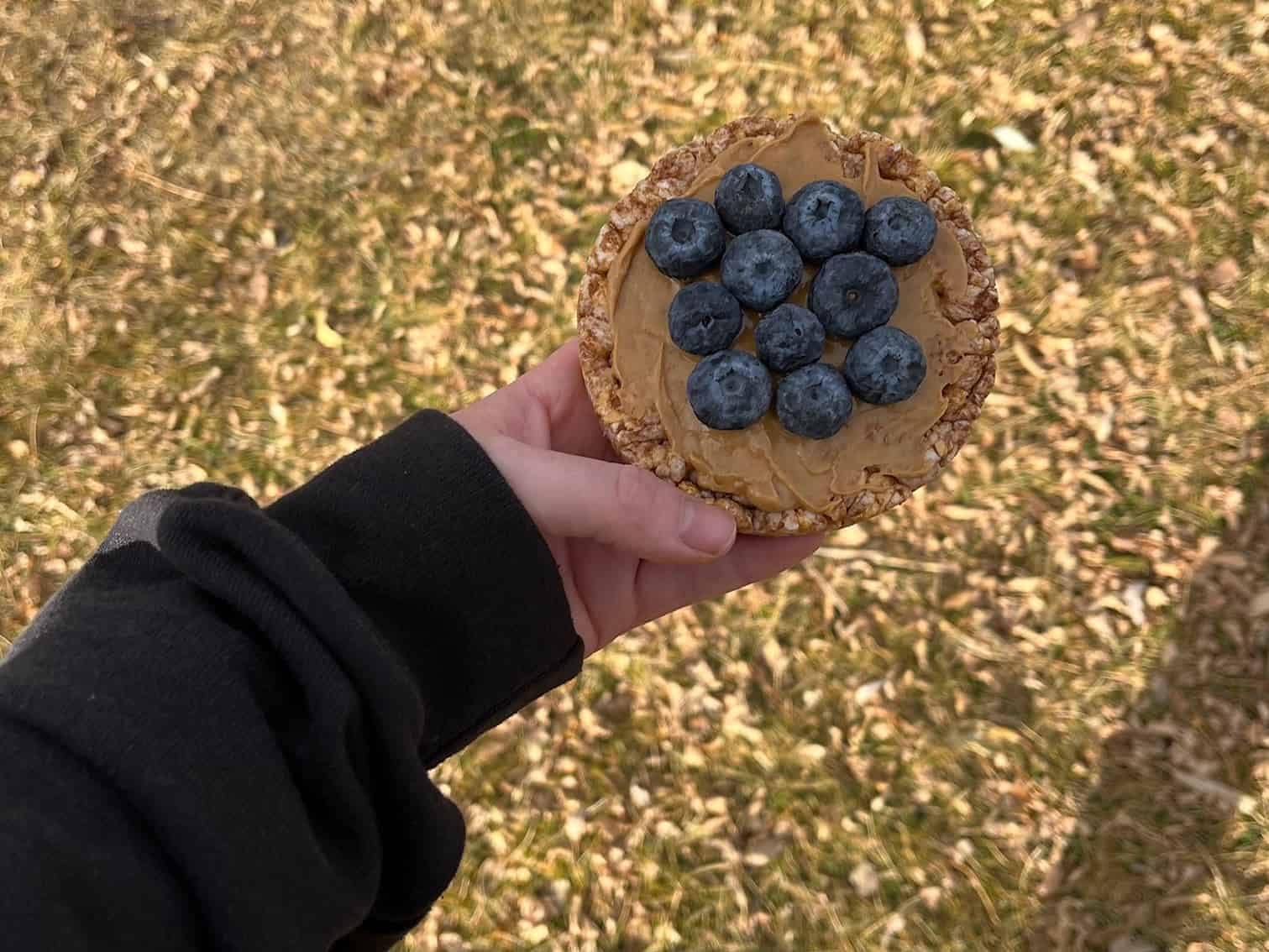 A woman's hand holds a rice cake topped with peanut butter and blueberries.