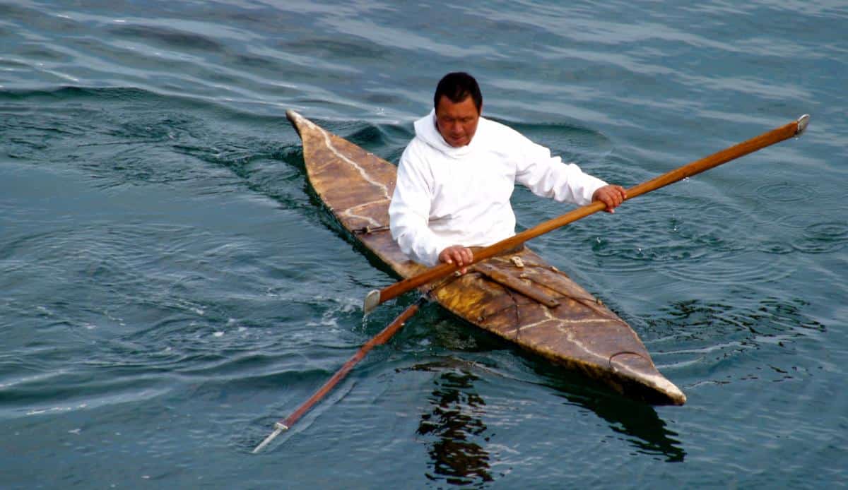 An inuk man holds a harpoon while sitting in his kayak.