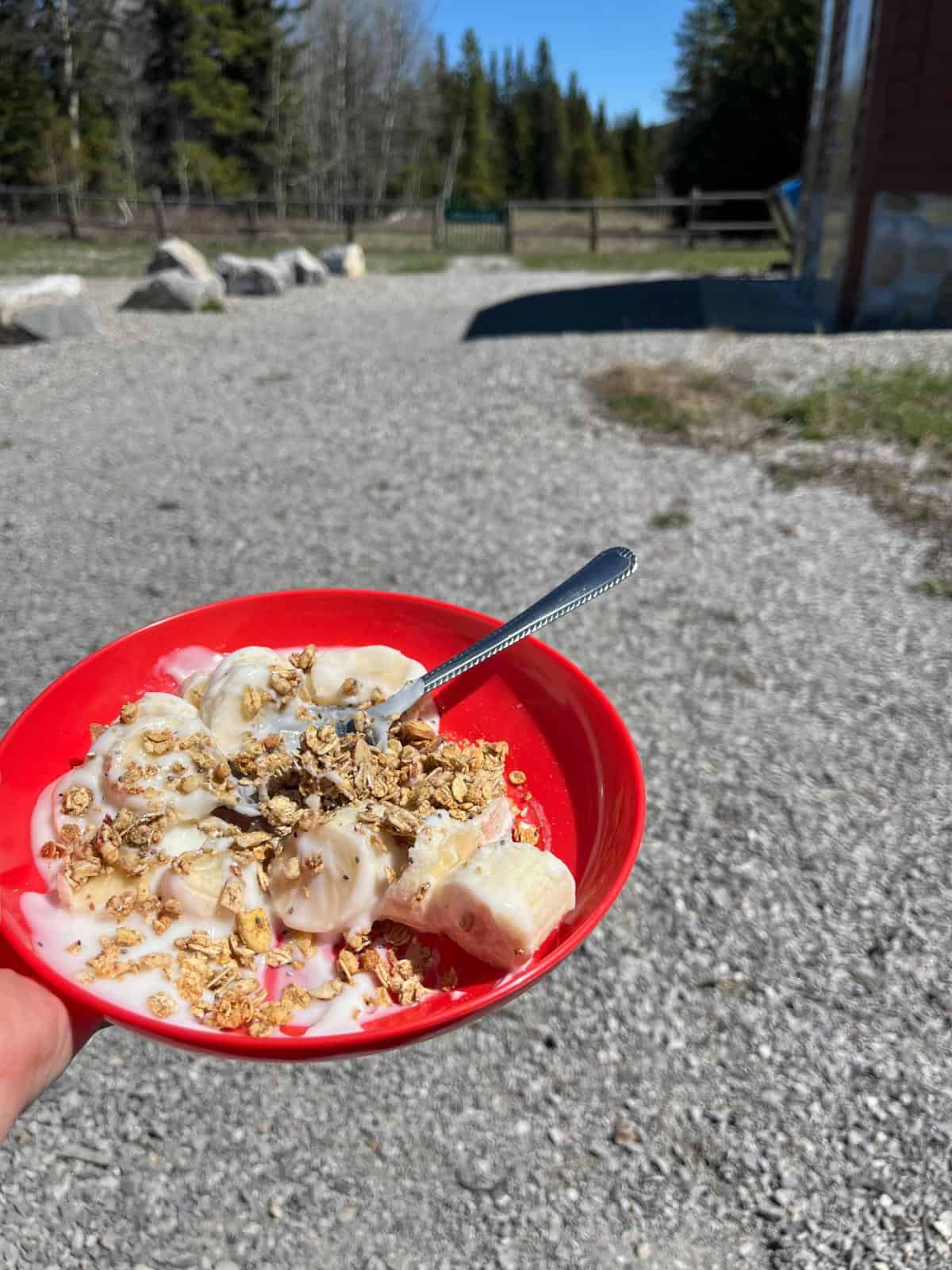 A red bowl full of yogurt, granola, and bananas. The picture is taken at a trail head and you can see trees and a fence in the background.