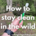 a woman's hand holds a green toothbrush up in front of a bunch of trees. the words "how to stay clean in the wild" are overlain on the image