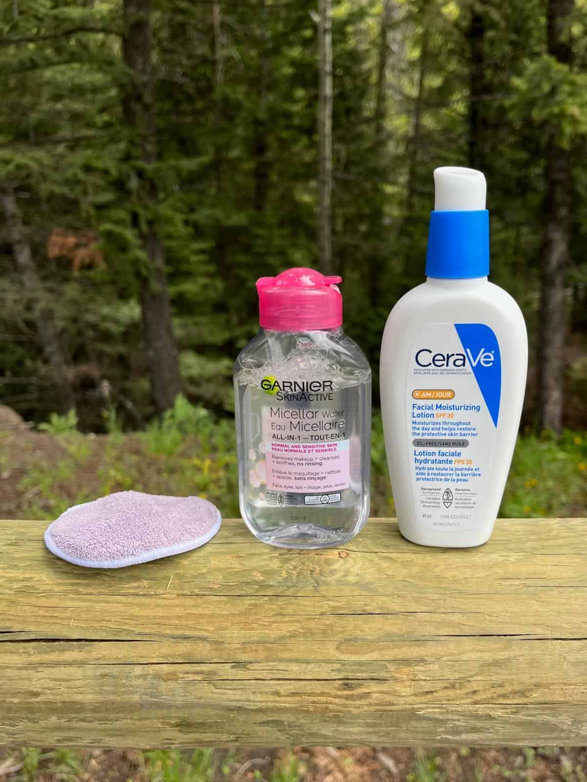 A cotton round, a bottle of micellar water, and a bottle of moisturizer with SPF sitting on a wooden fence