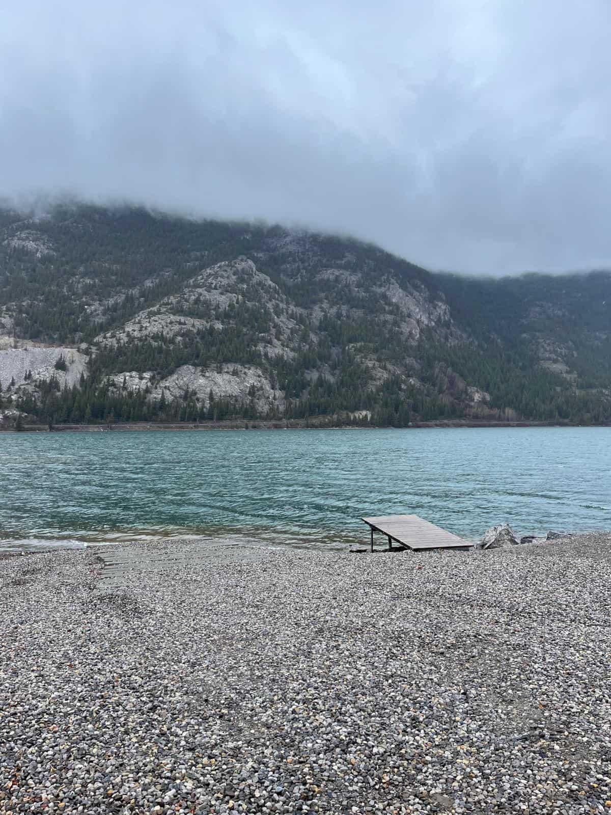 A lake with foggy hills in the background and dock leading out to bright blue water.