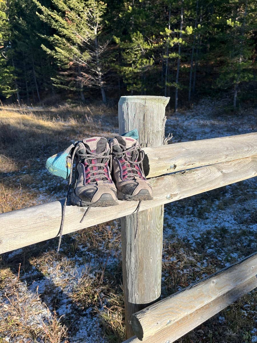 A pair of hiking boots rested on a thick wooden fence. A pair of wool socks sticks out of the boots.