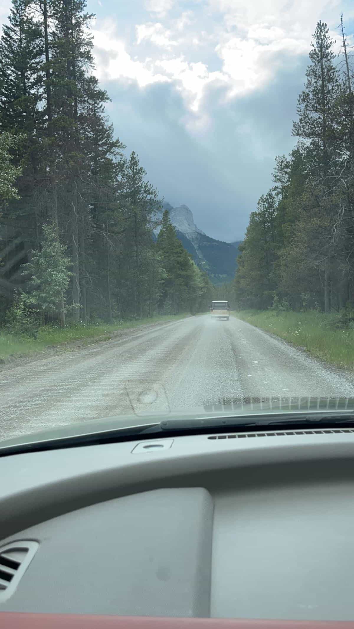 A vintage campervan drives down a gravel road with mountains in the background.