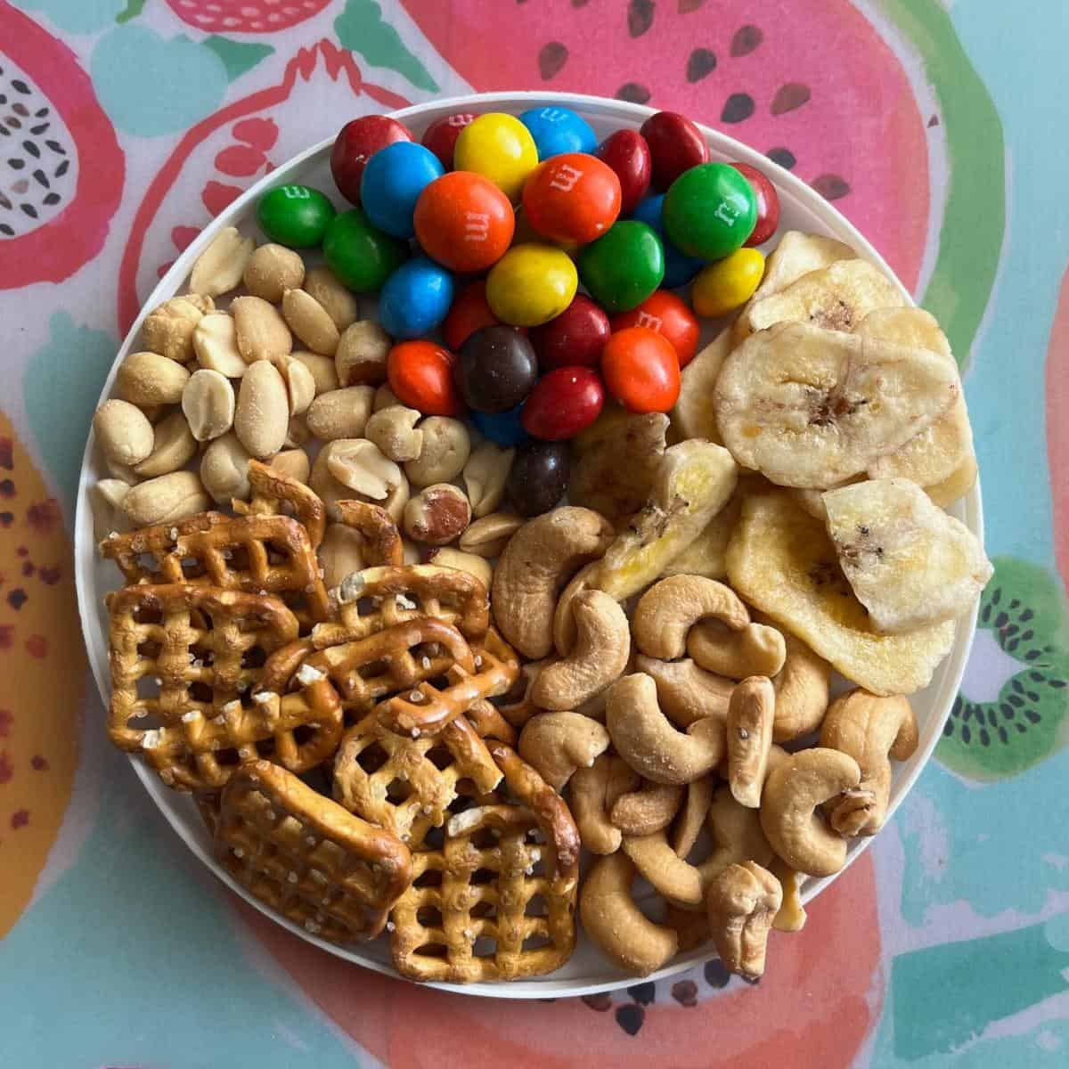 A plate with peanuts, pretzels, cashews, banana chips, and peanut butter M&M's.