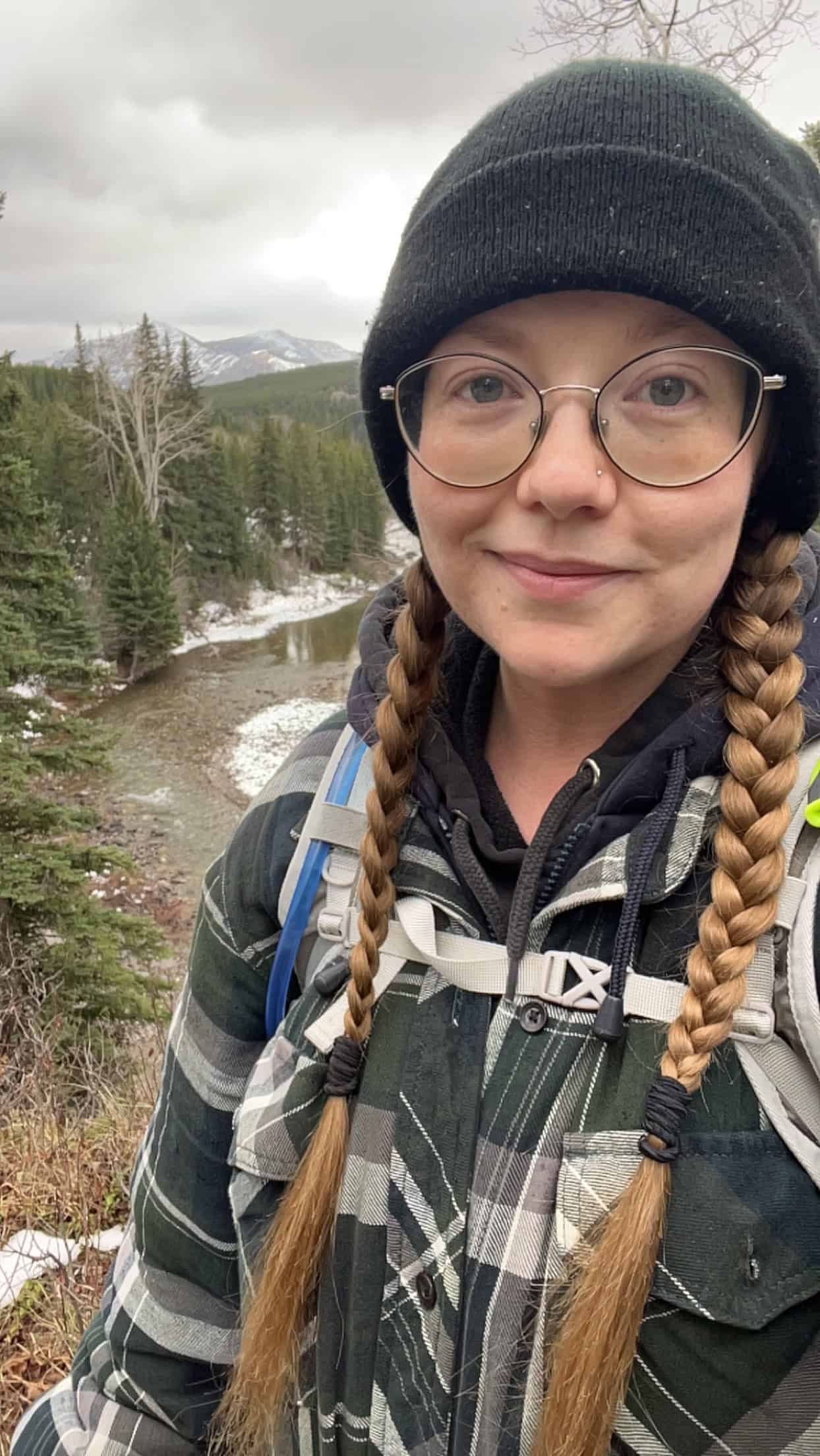 This is a picture of Danny. She is a Caucasian woman in her twenties. She is wearing a hiking backpack over top of a green flannel coat. Her blonde hair is in long braids that are covered by a black toque. She has big glasses. She stands in front of a snowy river with mountains in the background.