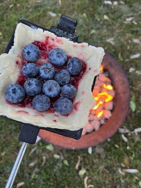 A pie iron over a fire. The pastry is missing its top, revealing a blueberry filling,