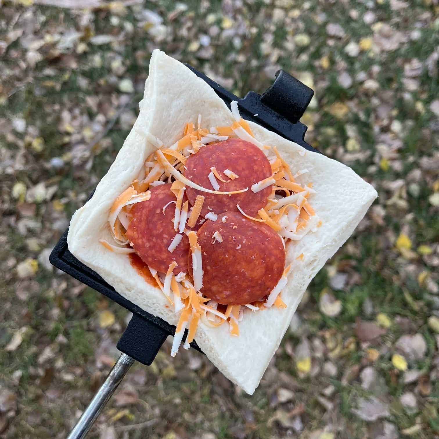 A pie iron pizza pocket before the crust is added on top. Sauce, cheese, and pepperoni are layered on a crust.