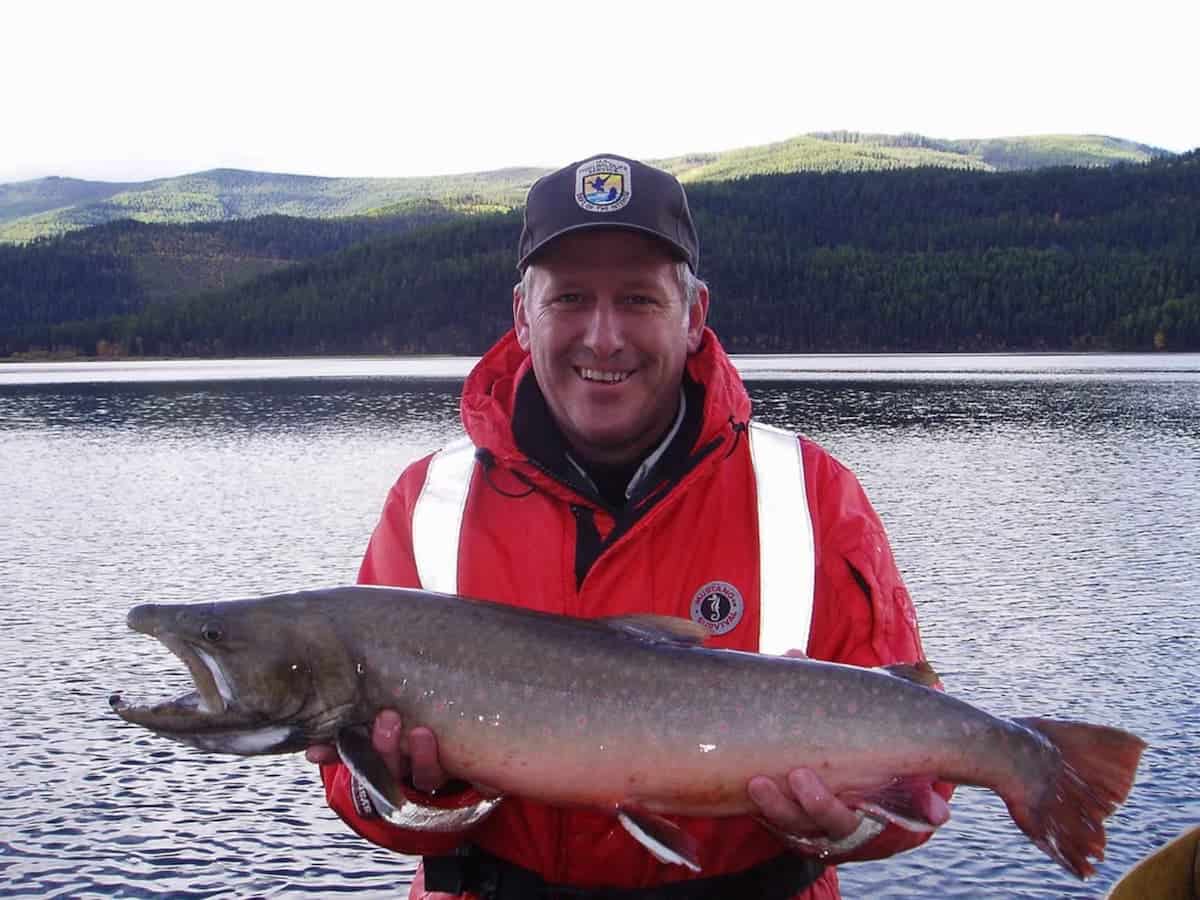 A man in a bright orange fishing jacket holds a large bull trout in front of a lake.